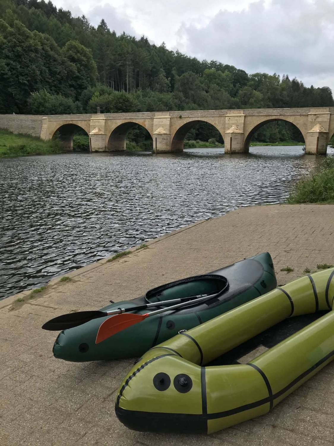 Deux packrafts gonflés posés sur une cale au bord de la rivière, devant un pont de pierre à arches