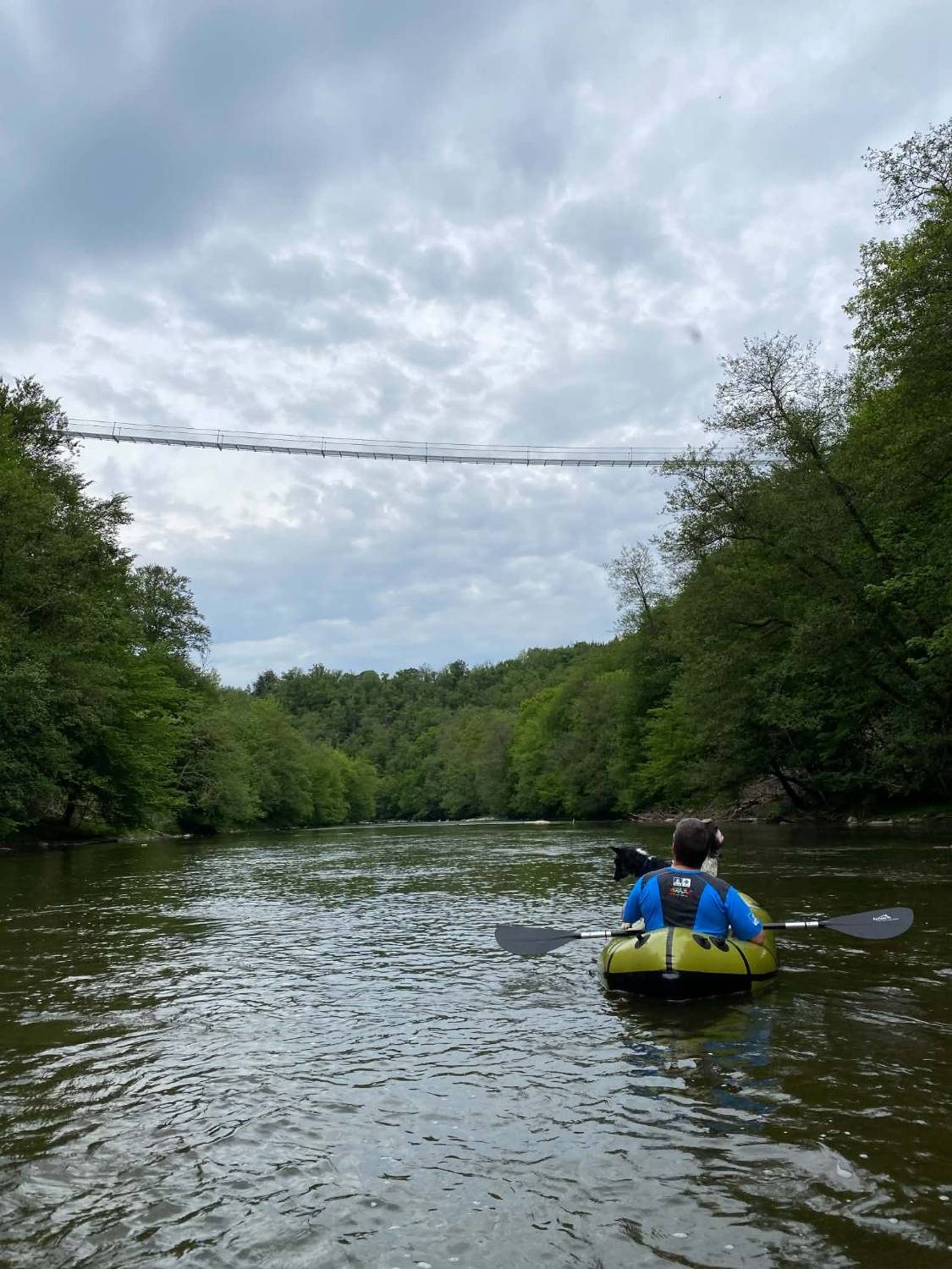 Un packraft glisse sur la Semois sous la passerelle suspendue de Sainte-Cécile, avec un chien à bord
